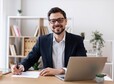 Smiling young Caucasian adult male in formal suit working at desk with laptop and papers. Professional, productive, and focused business setting showcasing modern office atmosphere.
